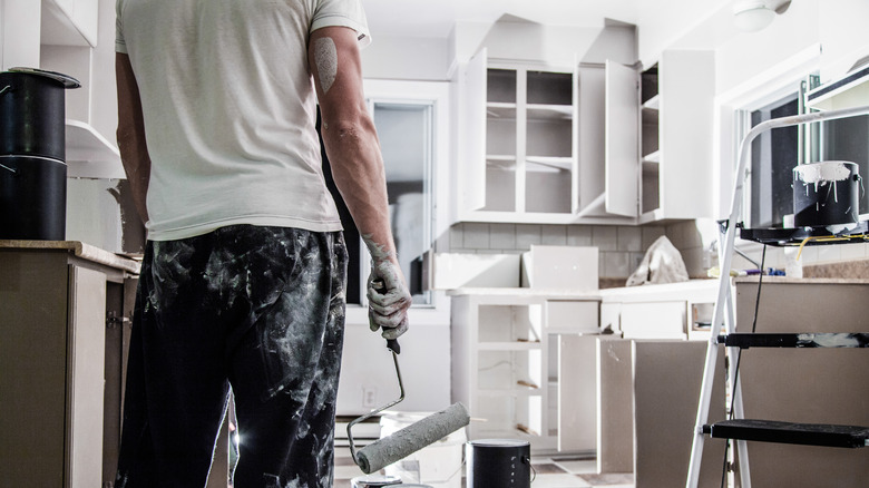 the back of a man holding a paint roller and covered in paint with an unfinished kitchen in the background